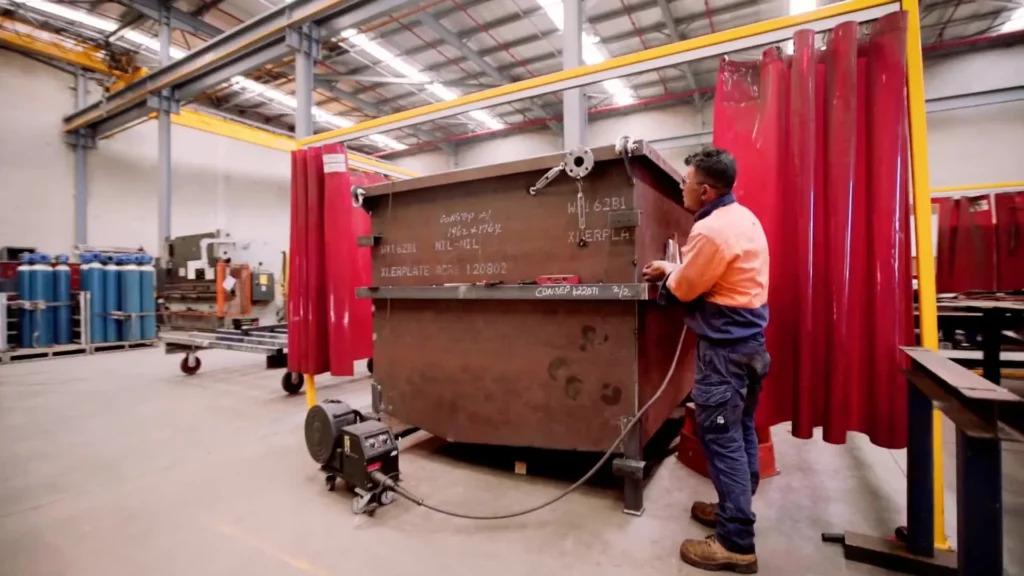 A person in an orange and blue work uniform operates machinery to handle a large, rusty metal container in an industrial workshop with high ceilings and red curtains. Various equipment is visible in the background.