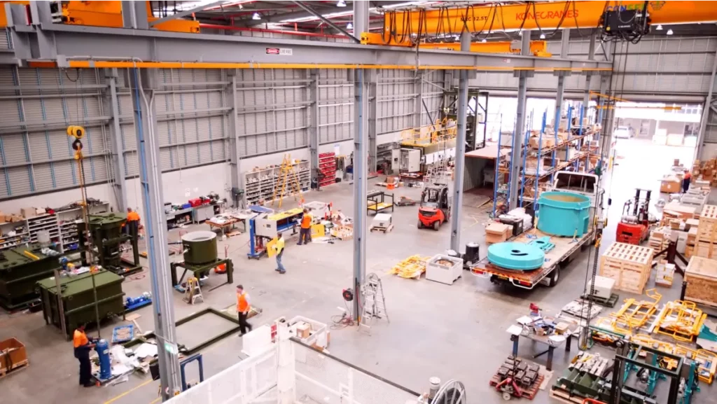 An industrial factory floor with workers operating and assembling heavy machinery. Various large equipment, tools, and materials are organized around the space. Overhead cranes and shelving units are visible. The area is well lit with high ceilings and metal beams.