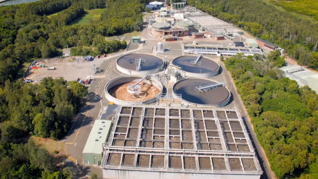 Aerial view of a large wastewater treatment plant surrounded by greenery. The facility consists of several circular clarifiers and rectangular sedimentation tanks. Buildings and infrastructure are spread throughout the site, with access roads running through it.