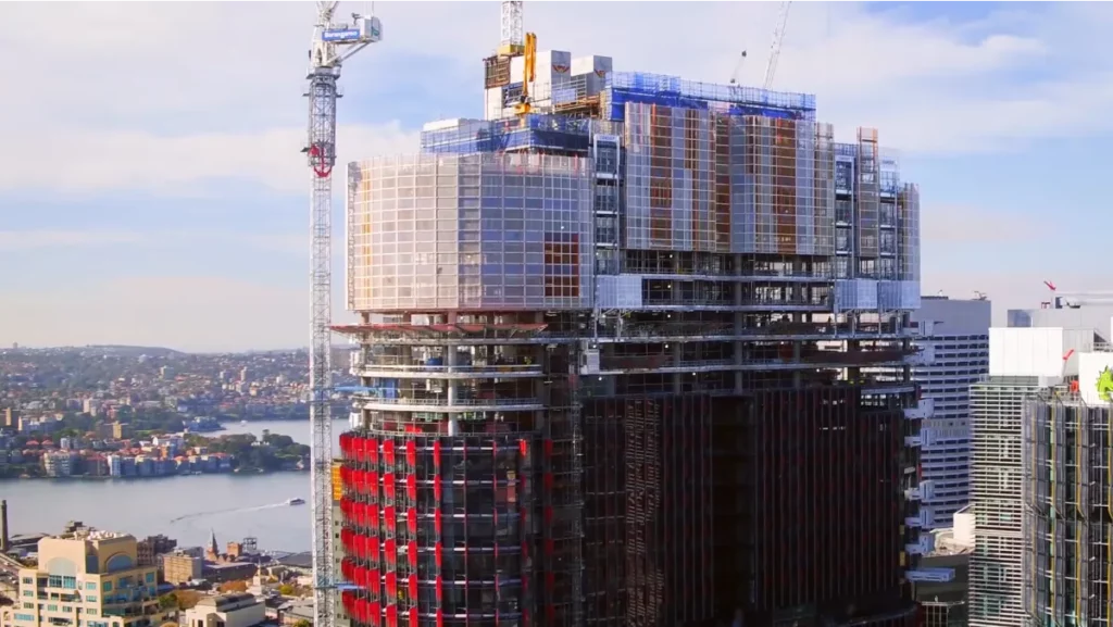 A tall, modern building under construction with cranes visible on its top. The structure features a mix of exposed and completed areas with glass windows and red accents. In the background, there is a body of water, other buildings, and a partly cloudy sky.