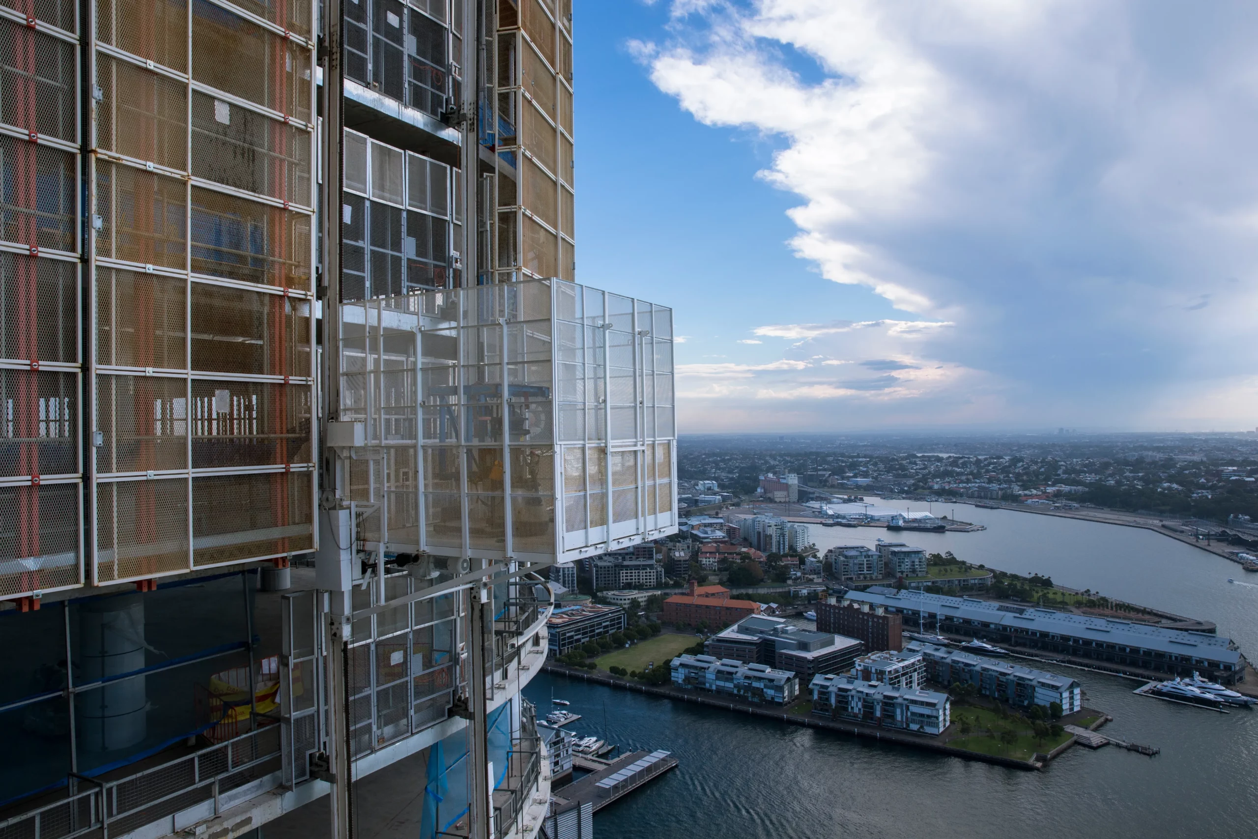 A construction elevator is seen ascending the side of a high-rise building under construction, overlooking a scenic waterfront cityscape. The sky is partly cloudy with blue sky peeking through, enhancing the view of surrounding buildings and water below.