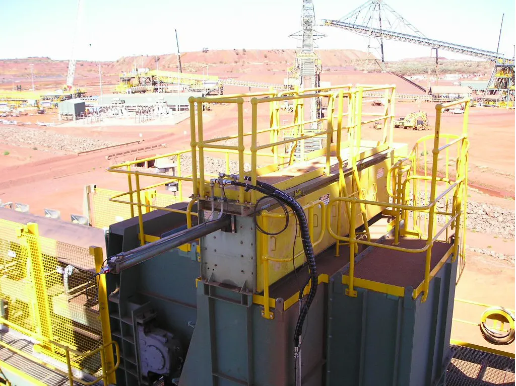 A large industrial machine with yellow safety railings and various cables is set against a backdrop of a mining facility in a desert-like area. The ground is reddish-brown and there are other pieces of heavy machinery and structures in the background.