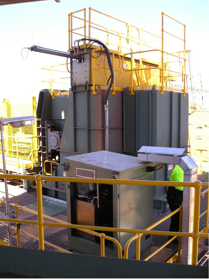 A large industrial machine with yellow safety railings, featuring multiple compartments and control boxes. There is a person in a neon green safety shirt and helmet interacting with the equipment. The background shows structural components and clear sky.