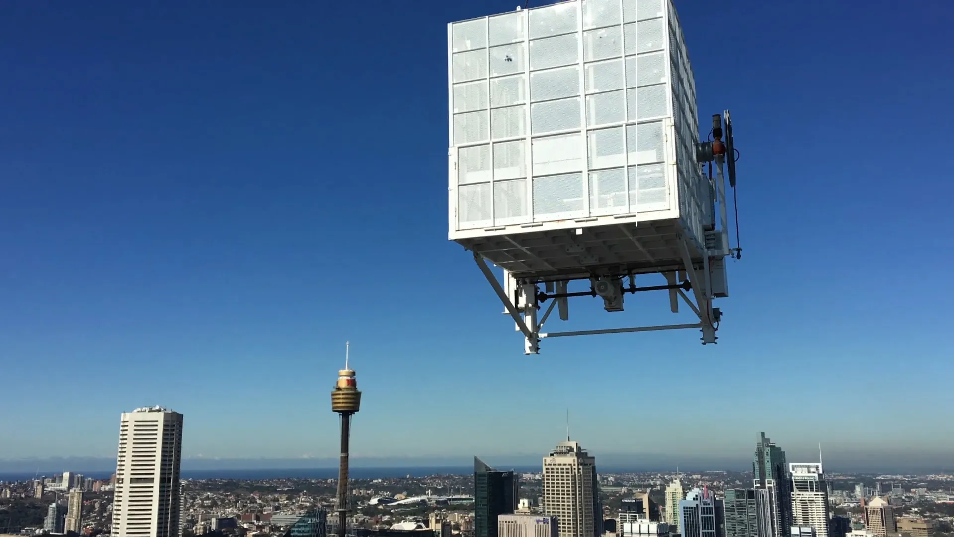 A large white structure, resembling a cube with glass-like panels, is suspended in mid-air against a clear blue sky. Below, a cityscape with tall buildings and a distinctive tower can be seen, extending to the horizon.