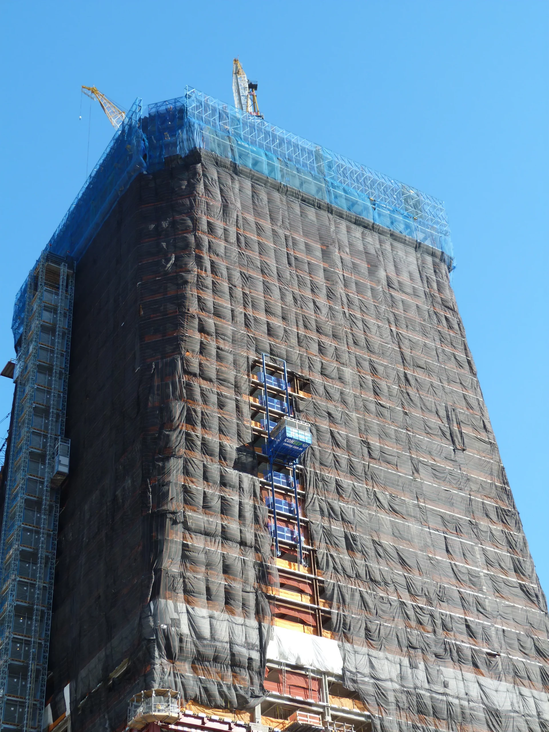 A high-rise building under construction, wrapped in black netting for safety, with visible crane and scaffolding at the top and sides. The clear blue sky serves as the backdrop.