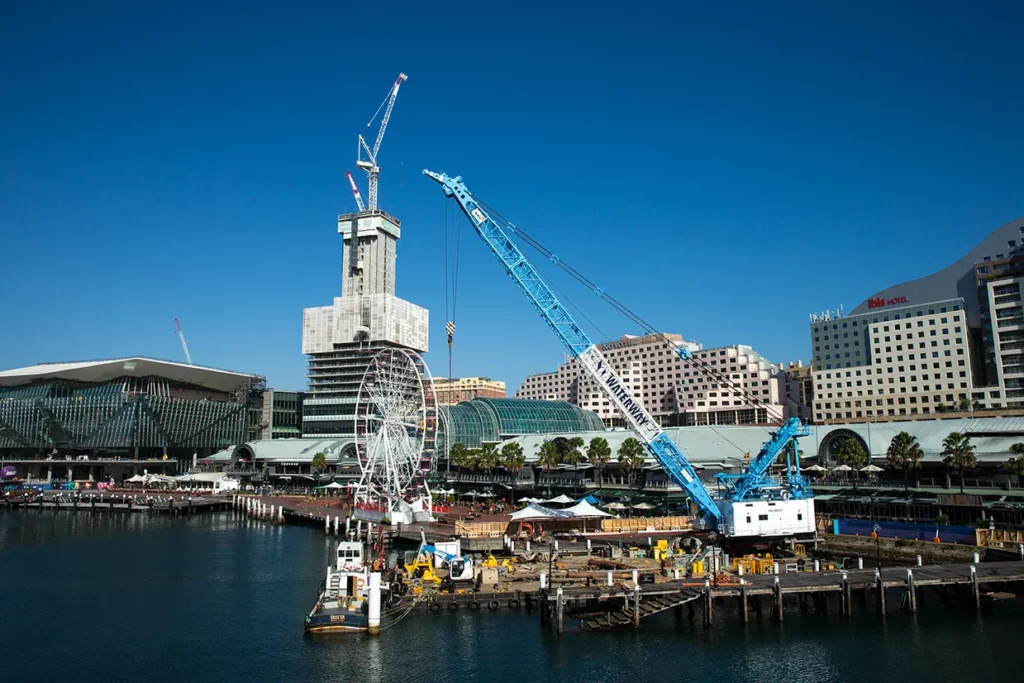 A waterfront construction site featuring a large blue crane, several buildings, and a tall structure under construction in the background. A Ferris wheel is also visible beside the water, and a clear blue sky stretches overhead.