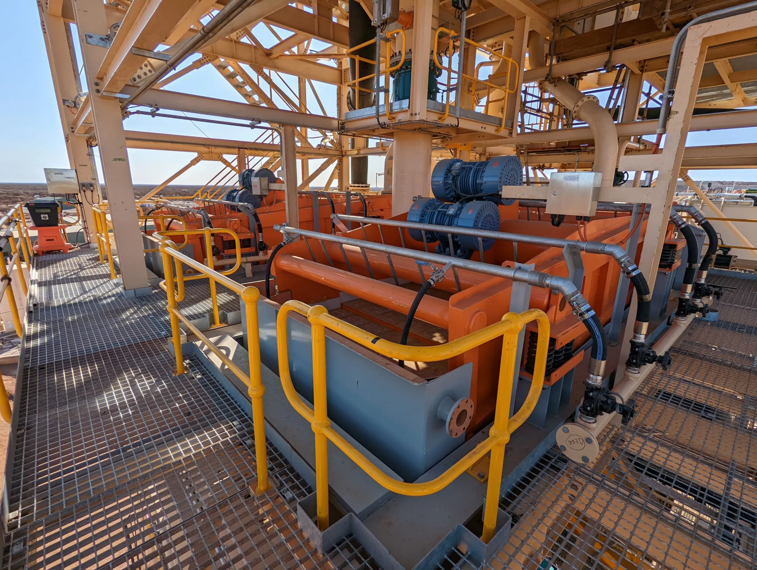 A section of an industrial facility featuring machinery with orange, blue, and yellow accents. The area has metal grating flooring, pipes, and railings. Bright natural light illuminates the structure, and an expansive view of the outdoors is visible in the background.