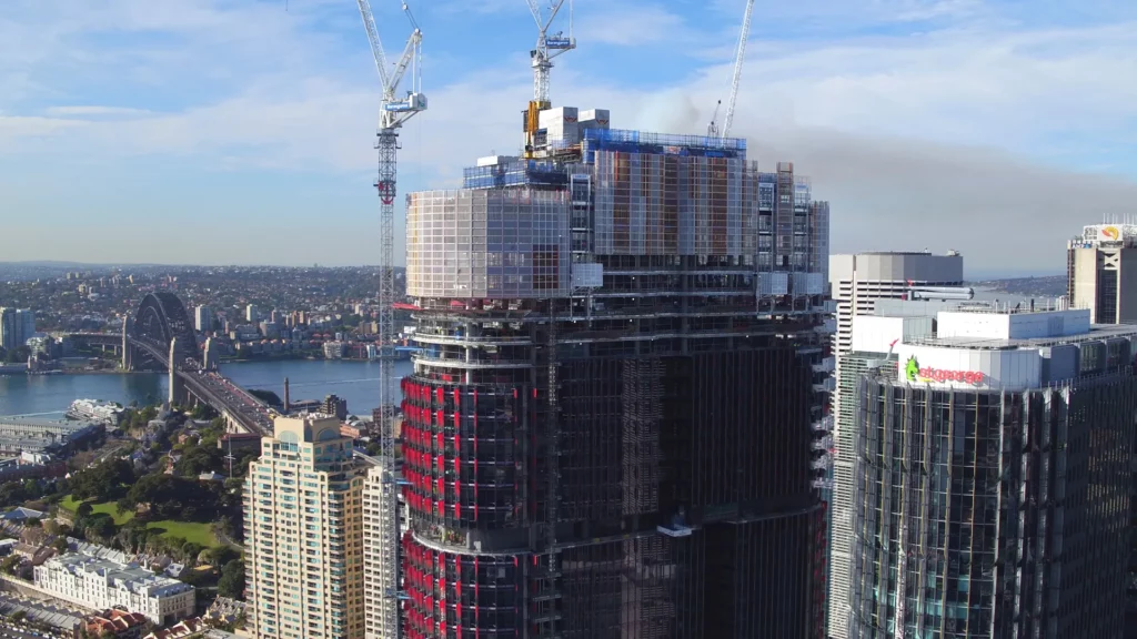 Aerial view of a construction site featuring a high-rise building under development in a city. Cranes are operating atop the structure. The cityscape includes buildings, a river, and a bridge in the background under a blue sky with light clouds.