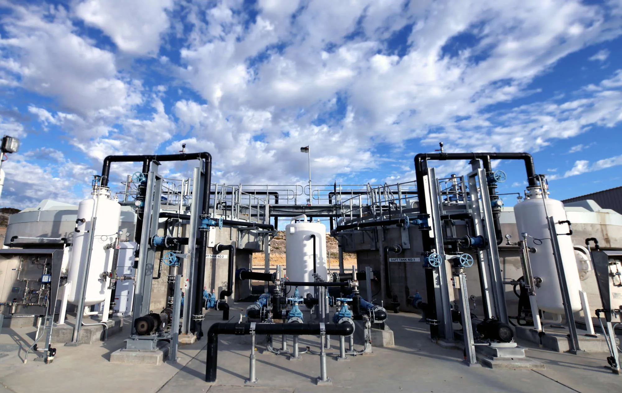 A large industrial water treatment facility under a partly cloudy blue sky. The plant features numerous pipes, tanks, and valves arranged in a complex network. The surrounding area is dry and appears to be in a desert or arid region.