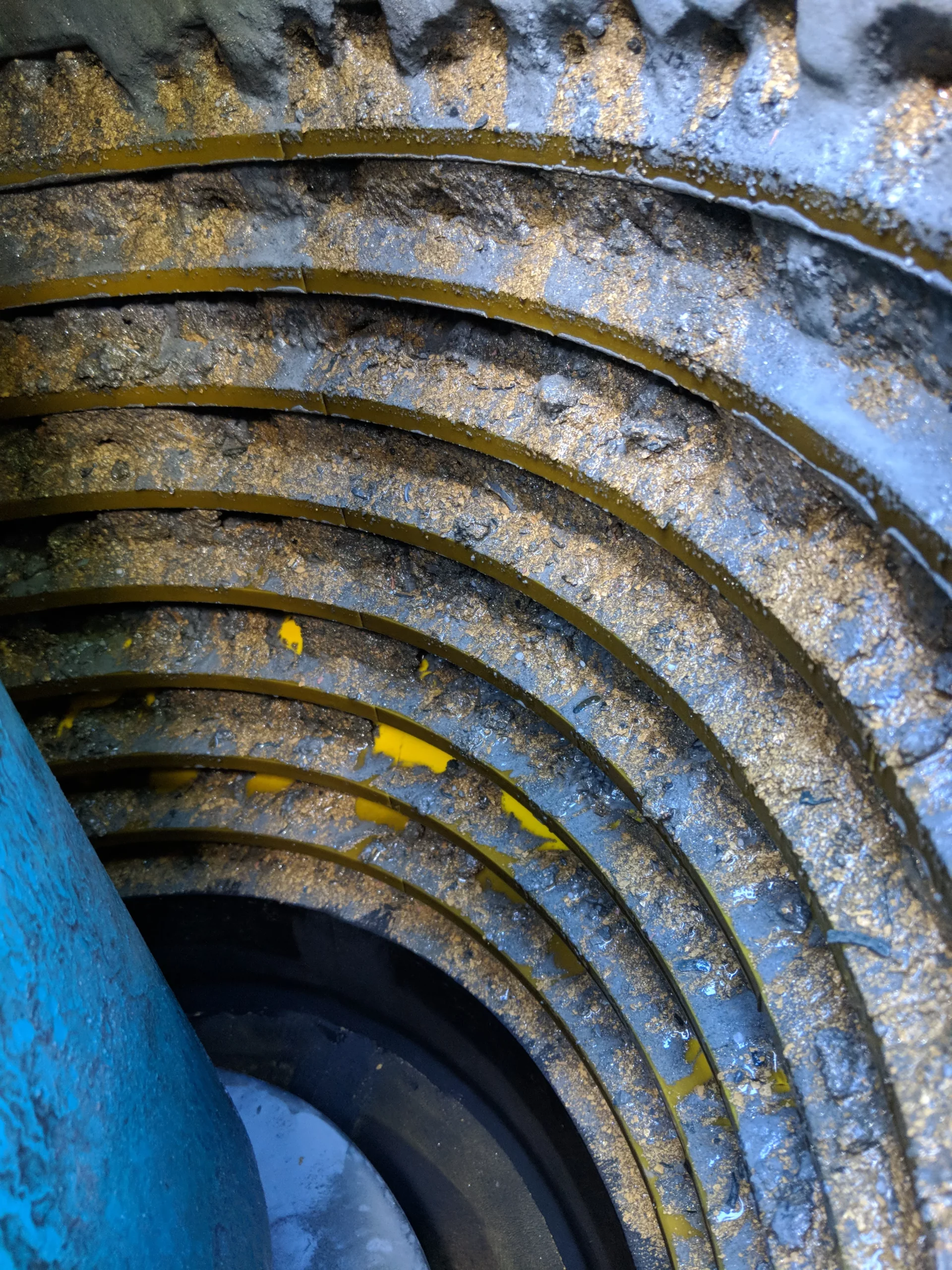 Close-up of a rusty, circular metal structure with rough, ridged edges and various shades of blue and yellow paint. The layers of rust and paint create a textured, weathered appearance.