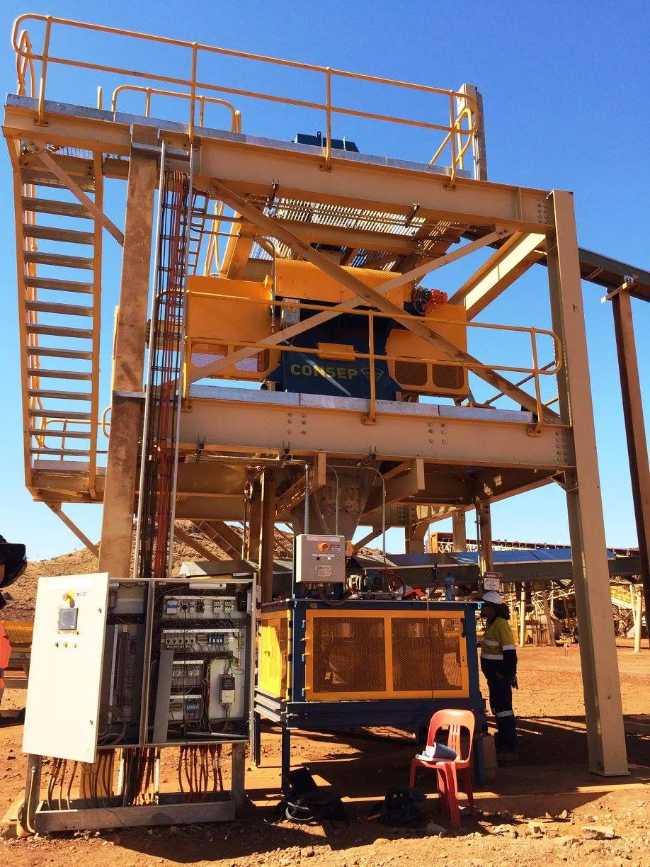 A large industrial mining machine stands outdoors in a deserted, sandy area. The structure includes a yellow and gray metal framework with various control panels and wiring. A worker wearing a protective helmet and vest is visible to the right of the equipment.