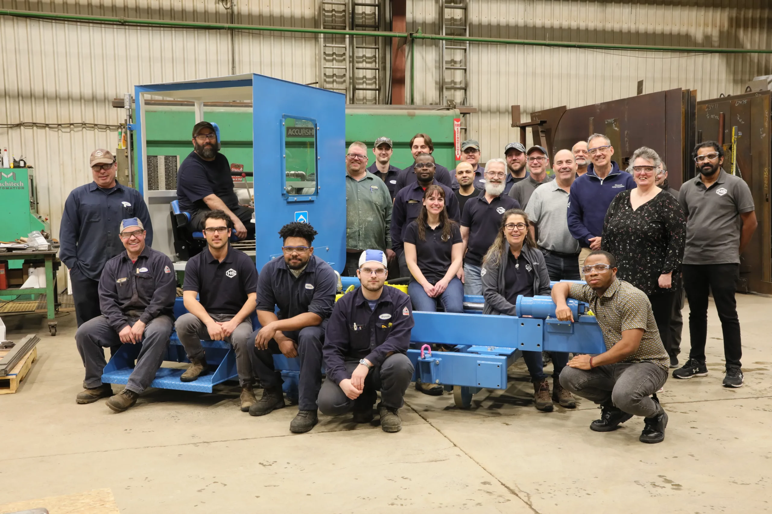 A group of people stand and sit around a large blue machine inside a warehouse. The group consists of both men and women, and they are dressed in a mix of work uniforms and casual clothes. The background shows various industrial equipment and tools.