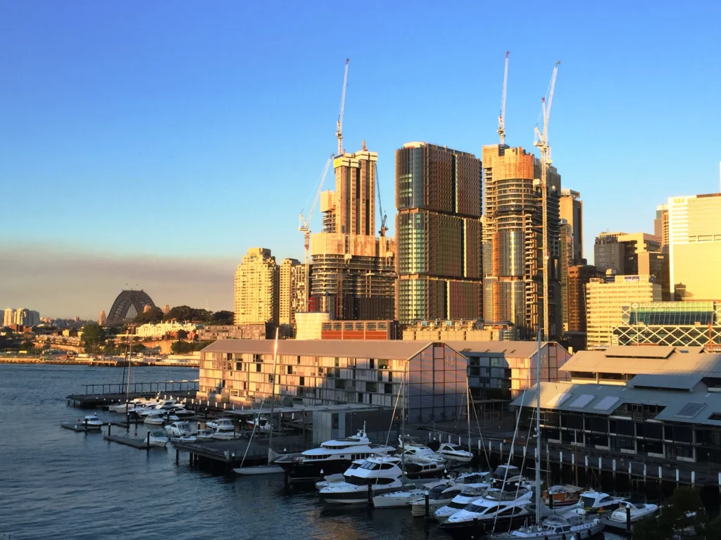 A vibrant cityscape of Sydney during sunset, featuring ongoing construction with cranes and tall buildings. The view includes moored boats in the harbor, illuminated by the golden light, with the Sydney Opera House visible in the background.