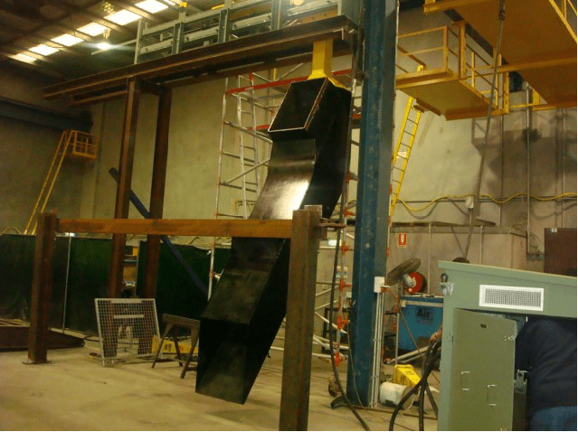 A large industrial workshop with a complex, curved black metal chute hanging from the ceiling. The chute is supported by wooden beams and extends downward. Surrounding the chute are various pieces of equipment, ladders, and yellow safety railings.