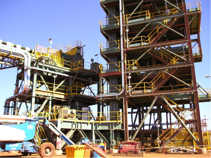 Image of a large industrial structure with multiple metal platforms and yellow railings. Various construction equipment and materials, such as a blue lift and a red toolbox, are scattered around the site. Clear blue sky in the background.