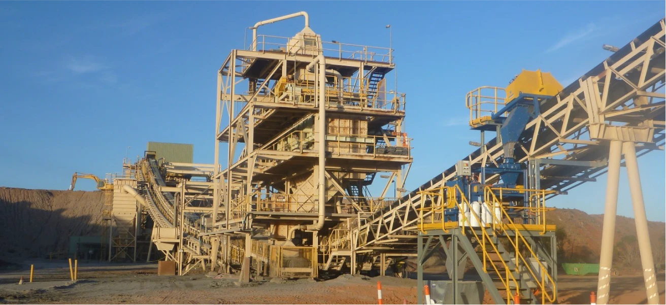 An industrial mining facility featuring large metal structures, conveyors, and machinery against a clear blue sky. The site includes multiple levels and walkways, with various pipes and equipment. The surrounding area is dusty and the ground is covered with gravel.