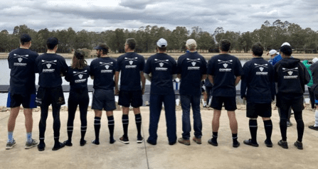 A group of ten individuals stand outdoors with their backs to the camera, wearing matching dark shirts with white lettering and graphics. They are aligned in a row on a concrete surface, facing a body of water and trees in the background.