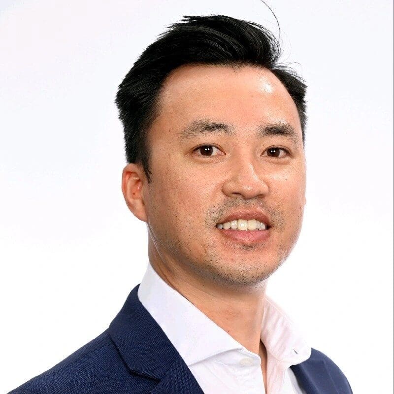 A man with short black hair and a slight smile is wearing a white shirt and a blue blazer. He is facing the camera against a plain white background.