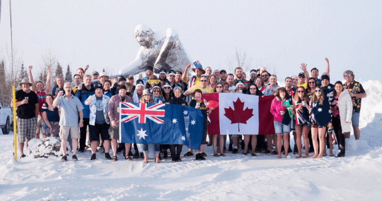 A large group of people dressed in casual winter attire are standing together in a snowy landscape, holding the Australian and Canadian flags. Behind them, a large snow sculpture in the shape of a human figure with wings is visible. The group is smiling and waving.