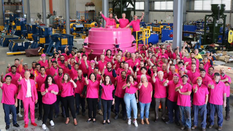 A large group of people wearing bright pink shirts pose together in an industrial warehouse setting, standing in front of a large pink industrial machine. They are smiling and giving thumbs up, showing a sense of unity and enthusiasm.