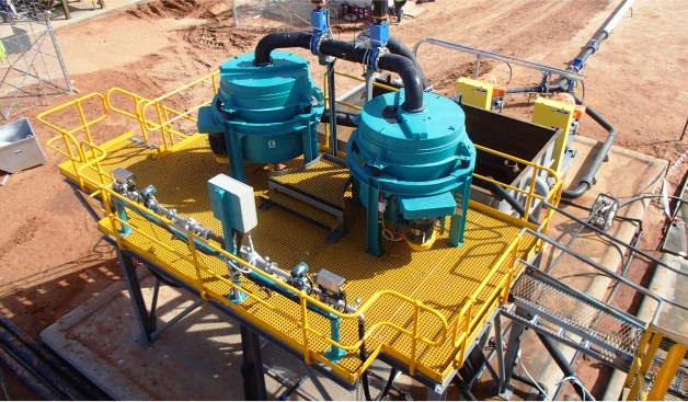 An overhead view of an industrial facility featuring two large, turquoise-colored pieces of machinery with black pipes. The equipment is installed on a yellow grated platform surrounded by safety railings. The surrounding area is a sandy construction site.