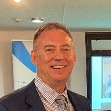 A man in a suit and tie smiles at the camera. He has short hair and stands in front of a conference room setting, with a banner and projector screen in the background.