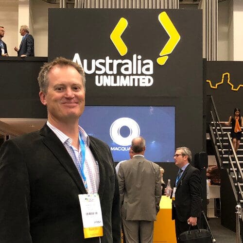 A man stands smiling in front of a booth with a sign that reads "Australia Unlimited." He is wearing a dark blazer and has a name tag around his neck. Behind him, several other people are conversing and interacting near the booth.