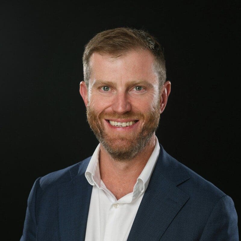 A bearded man wearing a white dress shirt and a blue suit jacket smiles against a solid black background.