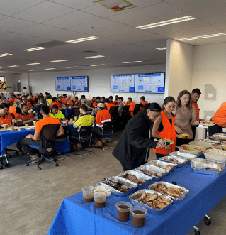 A large group of people, mostly wearing orange and yellow safety vests, are seated at tables having a meal in a brightly lit room. In the foreground, various foods and drinks are laid out buffet-style on blue tablecloths. Some individuals are serving themselves.