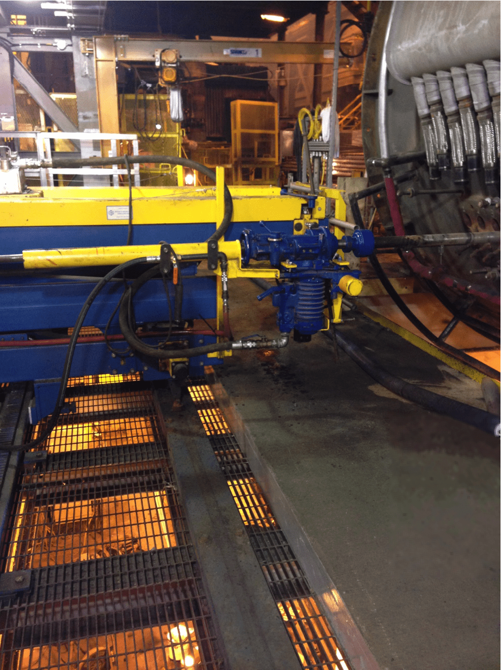 A factory floor with machinery, including pipes, a yellow railing, and various control systems. The equipment appears to be involved in an industrial process, with a grated metal walkway on the left and an area with multiple connection points on the right side.