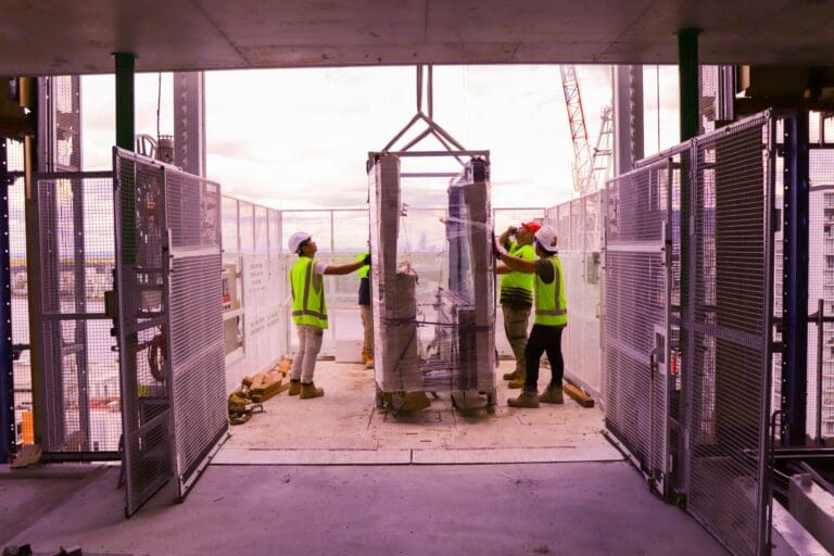 Three construction workers in safety gear guide a large, wrapped piece of equipment onto a building floor via a crane. They work on an open platform with safety gates, against a backdrop of a partly cloudy sky.