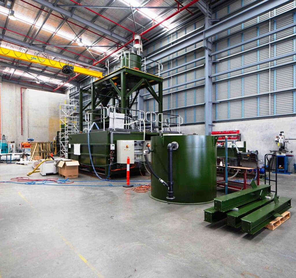 A large industrial machine with green metal frames and railings is situated in a spacious warehouse with high ceilings. Surrounding the machine are various tools, equipment, and materials including cables, boxes, and metallic components.