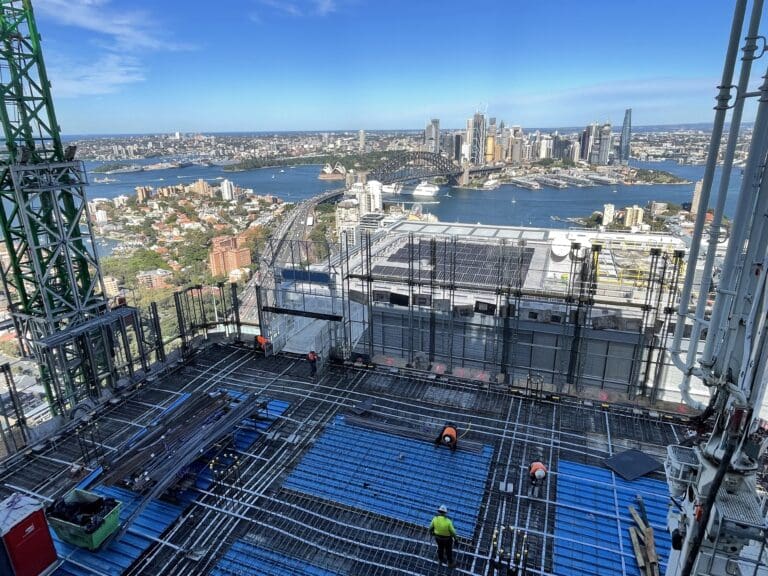 A high-angle view from a construction site atop a tall building. Workers in safety gear are on the rooftop, with blue panels and scaffolding visible. In the background, a cityscape with skyscrapers, water, and greenery is visible under a clear blue sky.