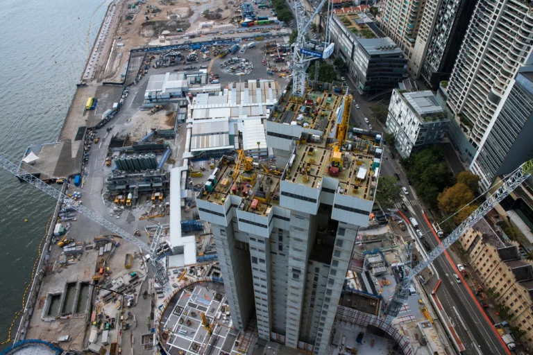 Aerial view of a construction site by a waterfront with cranes and numerous buildings in progress. Adjacent high-rise buildings line the street below, and various construction materials and machinery are spread around the site.