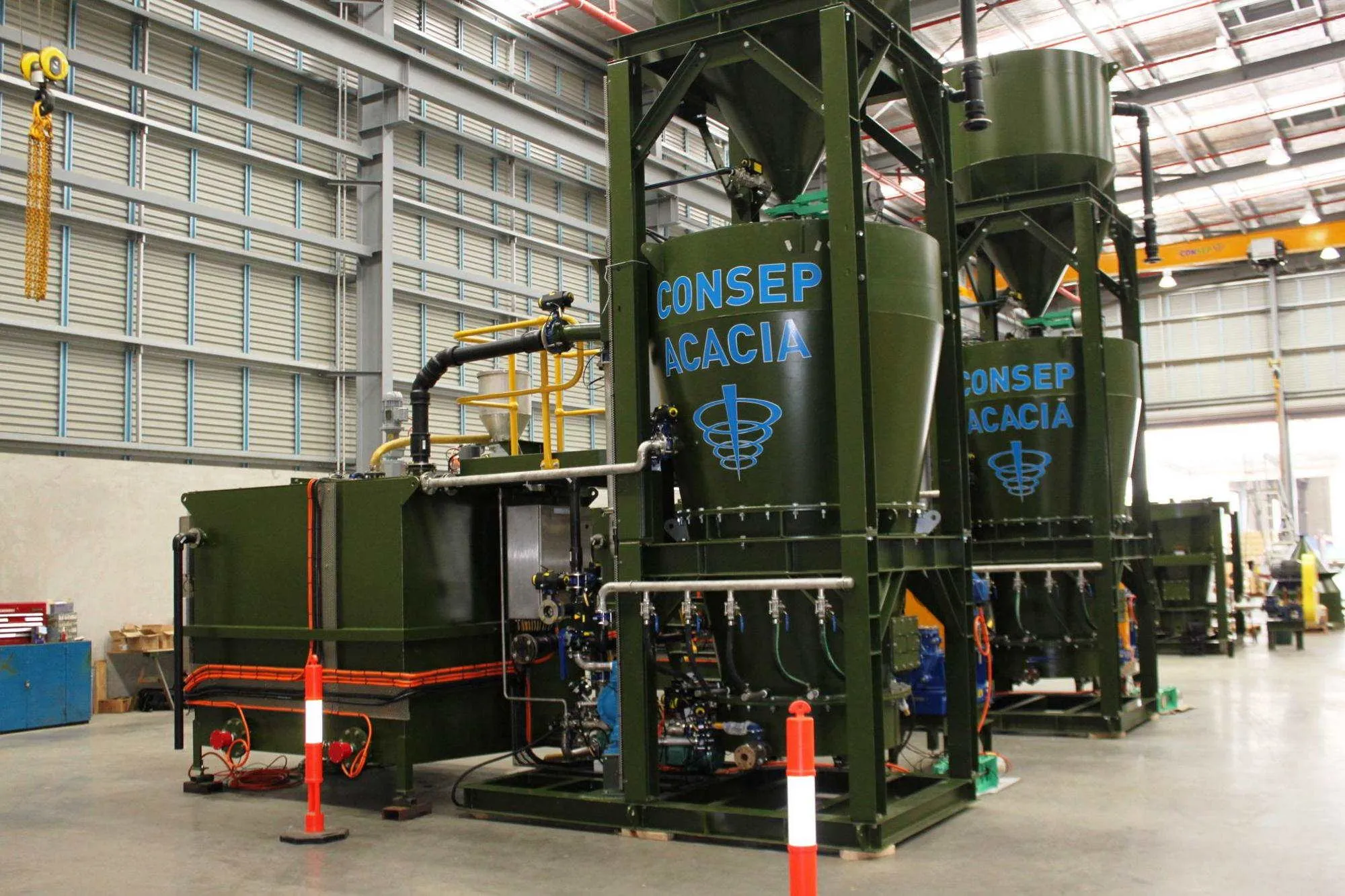 Industrial machinery setup within a large warehouse. The equipment features two tall, green tanks labeled "CONSEP ACACIA," connected with various pipes and components. The area is well-lit and partially enclosed by metal barriers.