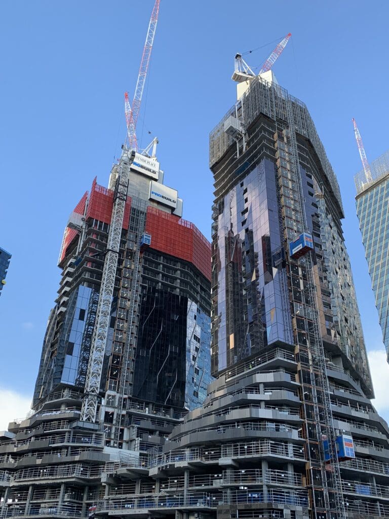 A cityscape showing two modern skyscrapers under construction. The buildings have glass facades and are surrounded by cranes. The sky is clear and blue, emphasizing the urban development.