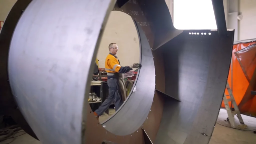 A worker in an orange safety jacket and black gloves is walking through a large, circular metal structure in a workshop with various tools and equipment in the background. The photo is taken from the inside of the metal structure, framing the worker in the center.