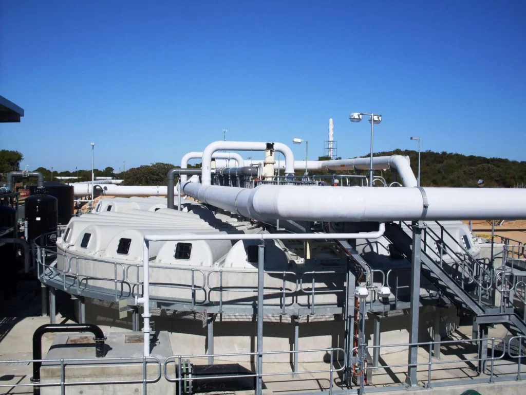 An industrial facility with large white pipes running horizontally above cylindrical tanks. The area is surrounded by railings and ladders for access. The background features a clear blue sky and sparse vegetation.