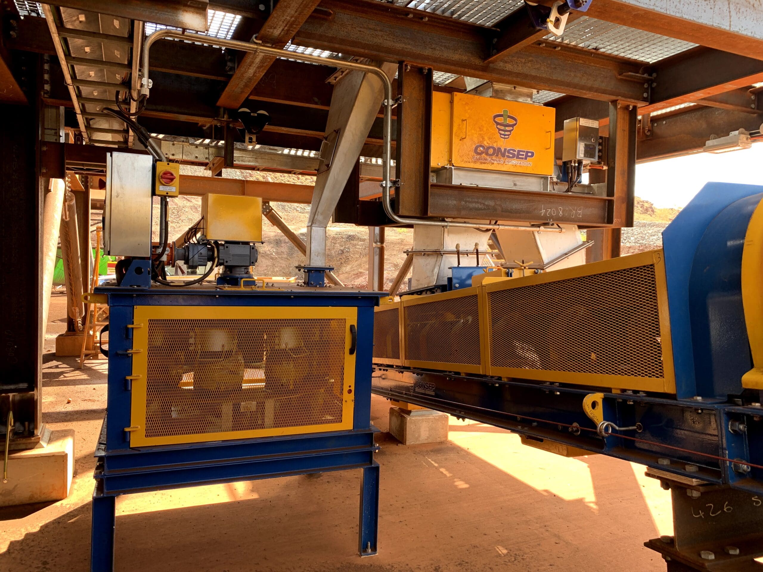 Industrial machinery setup under a metal structure, featuring conveyors and yellow mesh guards. Sunlight filters through, and various equipment components and a brand labeled "Consep" are visible. Dusty ground beneath the setup.