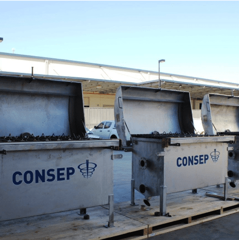 Three large, open metal CONSEP containers, placed on a wooden platform in an industrial outdoor setting. The sky is clear, and a white vehicle and part of a building with a corrugated roof are visible in the background.