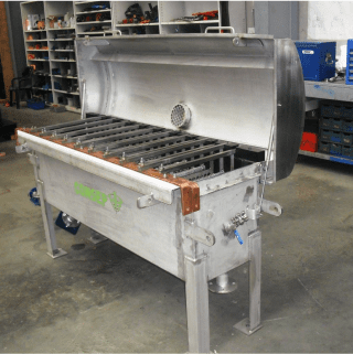 A large industrial stainless steel grill with a raised lid is situated in a workshop. The grill has metal grates and stands on sturdy legs. Various tools and equipment are visible in the background on shelves.