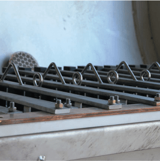 An industrial machine with rows of triangular metal brackets and bolts on a flat surface, with a circular, grid-like component in the background. The setting appears to be inside a factory or workshop.