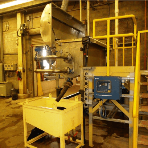 Industrial machinery in a factory, featuring a large metal container connected to pipes and various controls. A smaller yellow container is situated below, and there are steel stairs on the right side. The environment has an industrial, utilitarian feel.