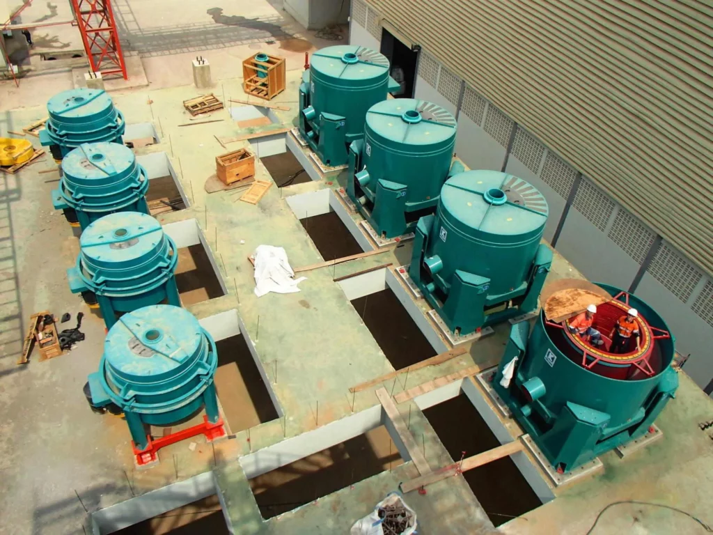 A construction site featuring multiple large, cylindrical, green industrial machines, possibly water pumps or filtration units, positioned over rectangular pits in the ground. Some of the equipment is partially installed, and various tools and materials are scattered around.