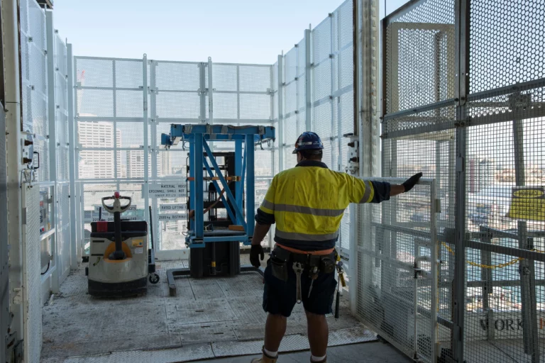 A construction worker in safety gear, including a hard hat and high-visibility clothing, signals with his hand on a construction site. The area is enclosed by metal fencing, and construction equipment such as a pallet jack and a lift are visible in the background.