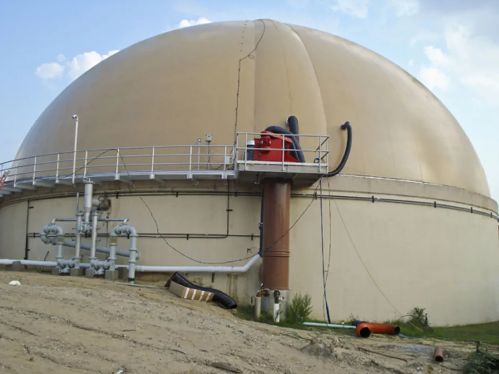 A large industrial dome structure stands at a worksite. The dome is beige and has a system of pipes and platforms attached to it. There are various pipes, hoses, and a red piece of machinery connected near the center of the dome. Dirt and debris are in the foreground.