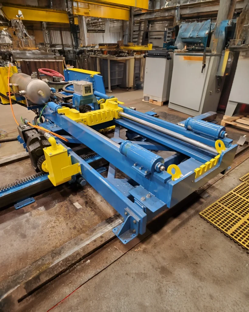 A blue and yellow industrial machine in a factory setting. The machine features rollers, motors, and various components, positioned on a concrete floor with industrial equipment visible in the background.