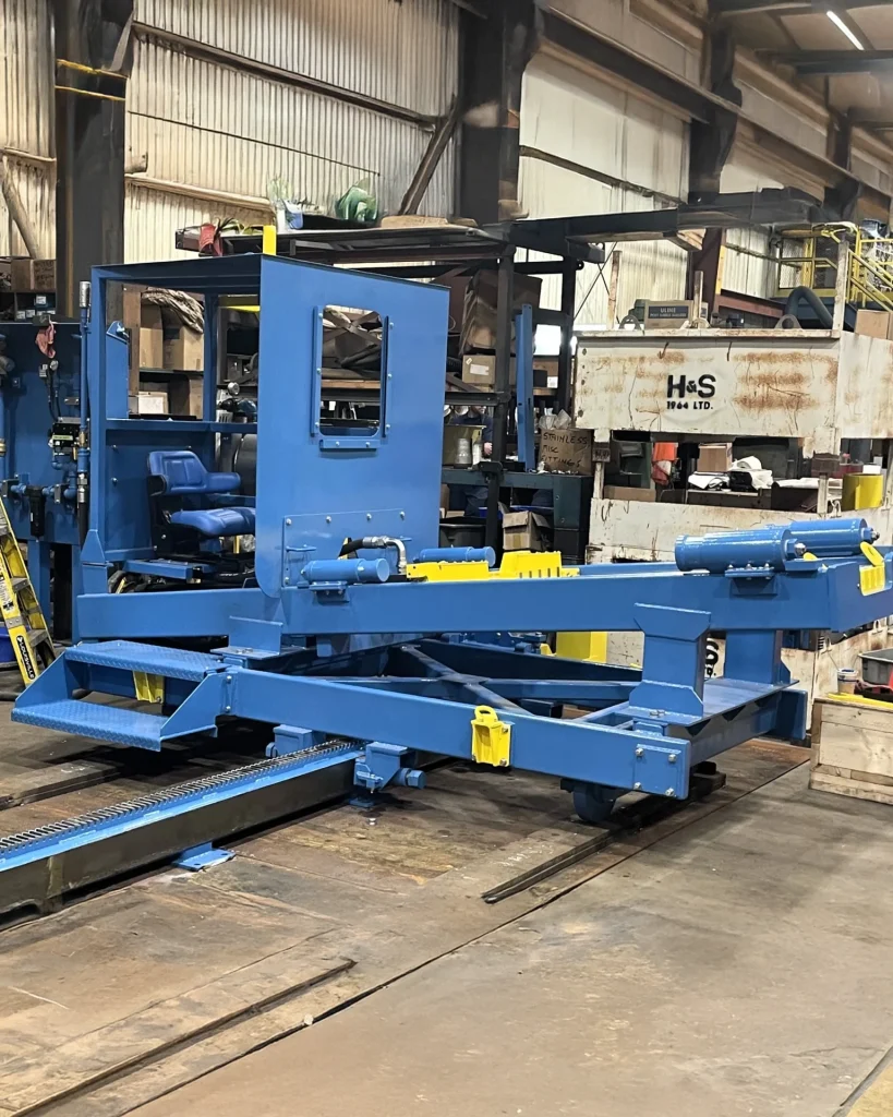 A blue industrial machine with a complex framework and yellow accents positioned inside a metal fabrication workshop. The surrounding environment includes shelves with tools and equipment, and metal beams in the background.