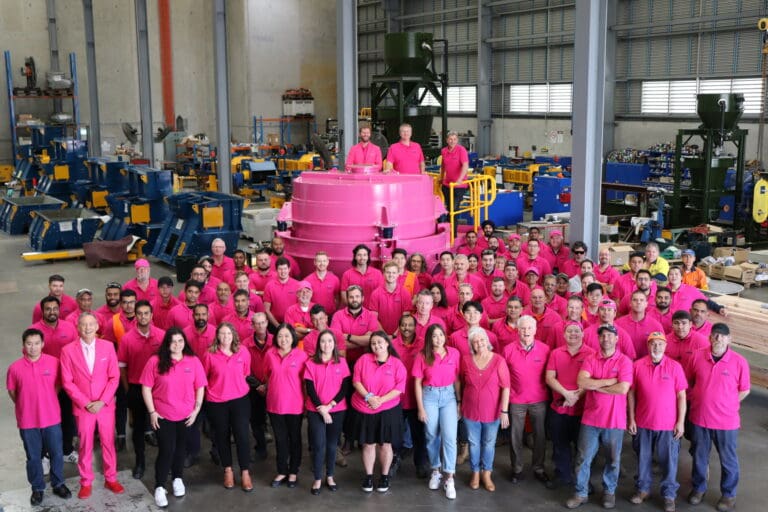 A large group of people, mostly wearing bright pink shirts, pose together inside an industrial warehouse. In the center is a large, pink machine. Workshop equipment and machinery are visible in the background.