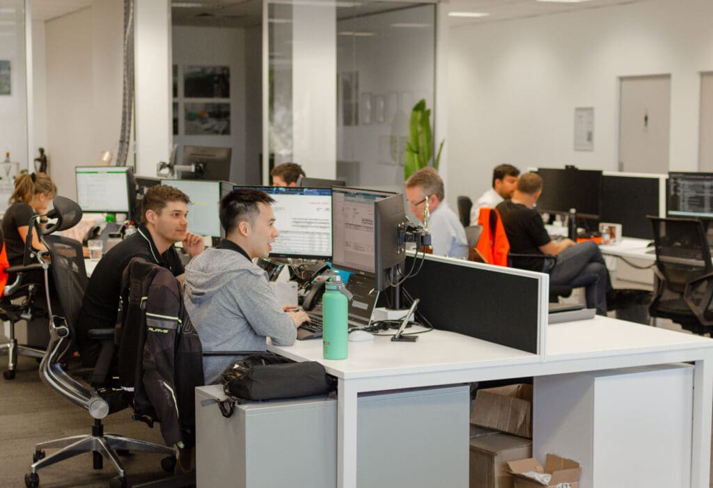 People working in a modern office with desks and computers. Several individuals are focused on their screens, while others engage in discussions. An open layout with white walls and natural lighting is visible. A green water bottle is on a desk.